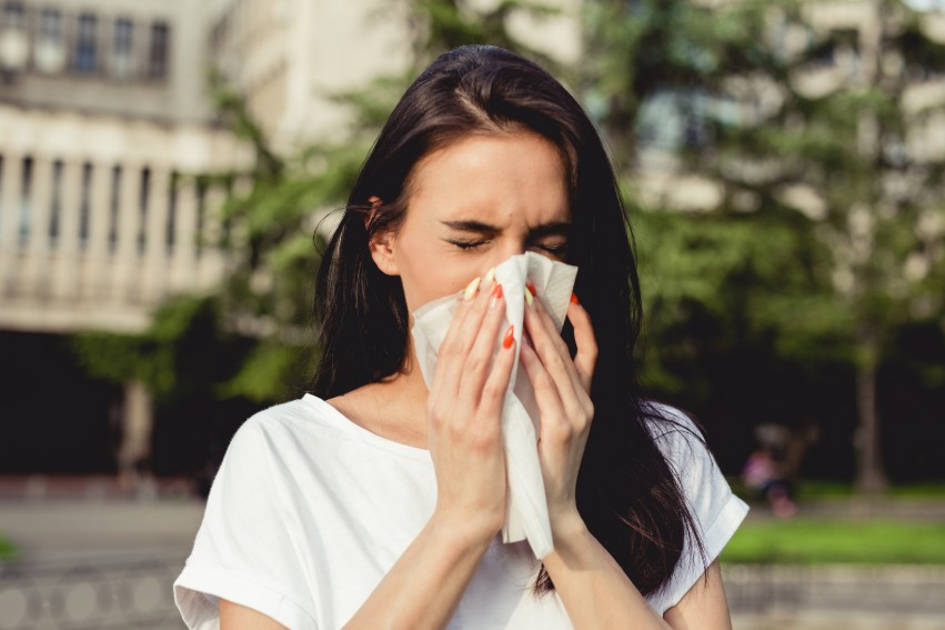 Woman outdoors with eyes closed, sneezing into a tissue while wearing a white shirt, with blurred city buildings and trees in the background. Woman outdoors with eyes closed, sneezing into a tissue while wearing a white shirt, with blurred city buildings and trees in the background.