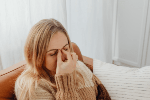 Woman sitting on a sofa indoors, eyes closed and pinching the bridge of her nose, appearing tired and experiencing sinus discomfort.