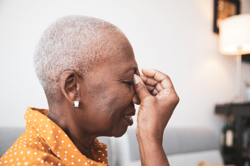 Older woman with short gray hair in an orange polka dot shirt gently pinches the bridge of her nose in a softly lit room.